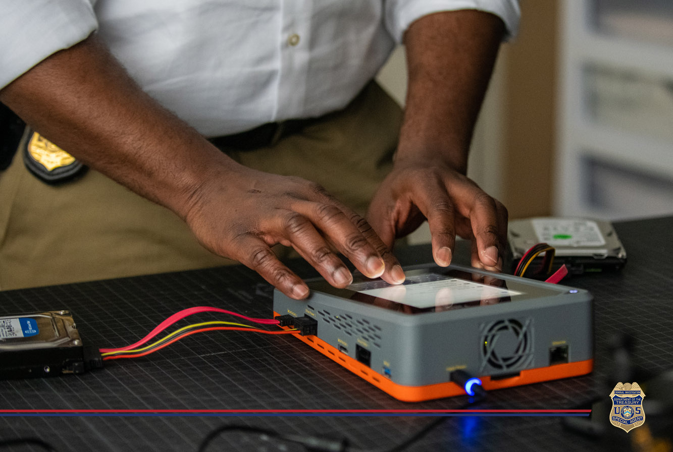 A forensic analyst in a white shirt uses a digital device to examine data, with focus on his hands manipulating the device on a desk filled with various electronic components.